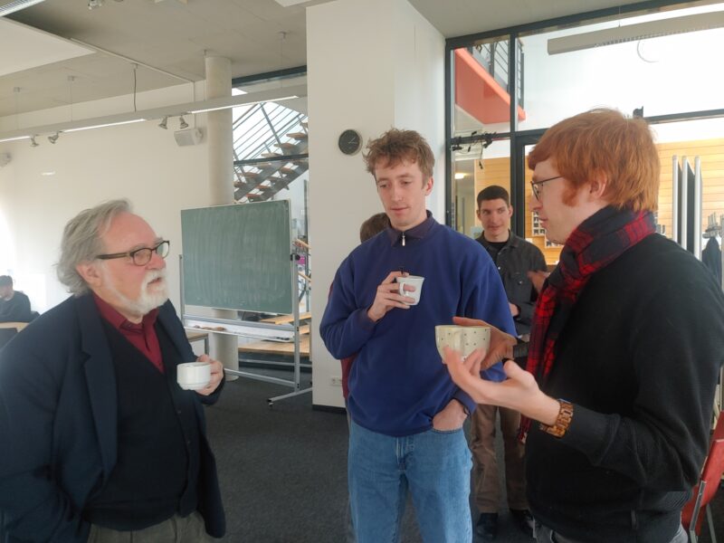 Three people standing and discussing holding pots of tea in hand. In the back one person just looking good.