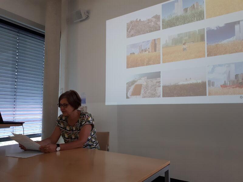Kirsten Twelbeck, sitting in front of the auditorium. In the background, a collage of the wheat field by Agnes Denes is projected onto the wall with the beamer
