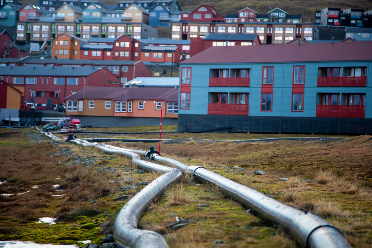 Pipelines can be seen running from the centre of the picture, making their way to a village in the background. The pipelines lie on a meadow.