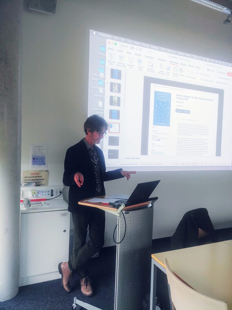 Person standing in front of lectern in the action of presenting. He is looking at his laptop and pointing his fingers to the horizontal sides.